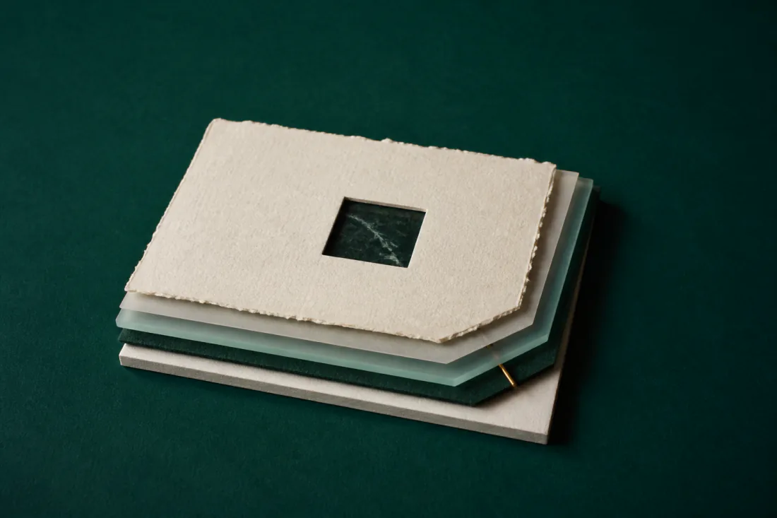 A stack of sheets — handmade paper, vellum, frosted glass, felt and board — fanned slightly apart, with a small square window cut through the top sheet