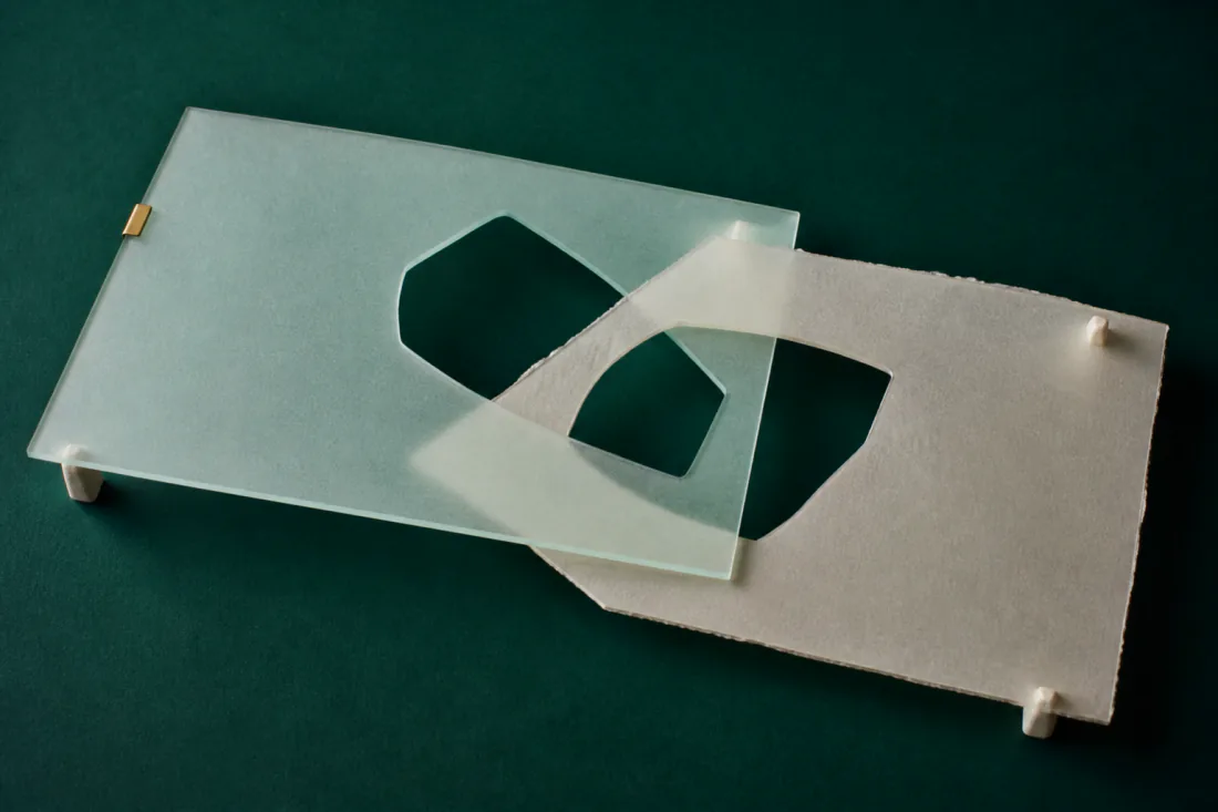 A sheet of frosted glass and a sheet of handmade paper, each cut with an opening, laid across one another so the two openings overlap to form a third shape